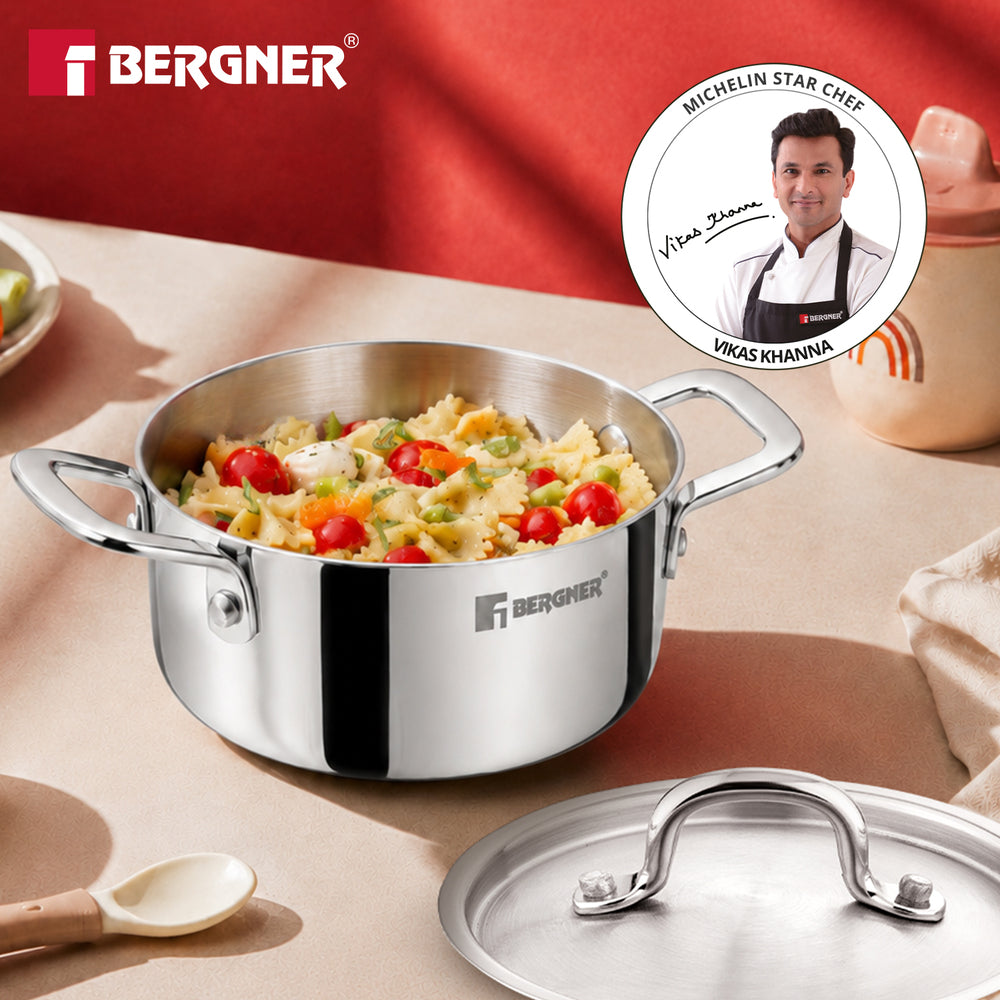 A Bergner stainless steel saucepan filled with pasta and cherry tomatoes, shown on a beige tabletop with its matching lid placed in front. A red Bergner backdrop and a circular Michelin Star Chef badge are visible in the scene.