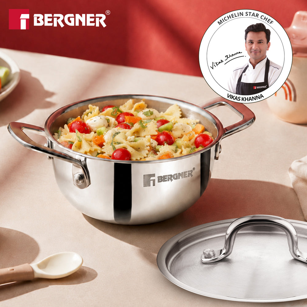 A stainless steel Bergner saucepan filled with pasta and vegetables, shown on a beige tabletop with its lid placed beside it. The Bergner logo appears in the top left, and a circular Michelin Star Chef badge featuring Vita Szepe and Vikas Khanna is shown in the top right.