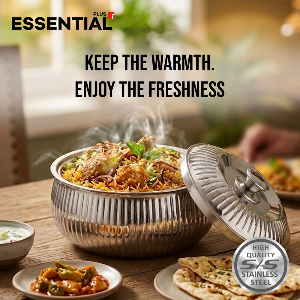 A stainless steel tandoor-style oven with its lid propped open, showing cooked food inside and steam rising. The scene is set on a wooden table with side dishes and a blurred dining background, with text reading 'ESSENTIAL' and 'KEEP THE WARMTH.